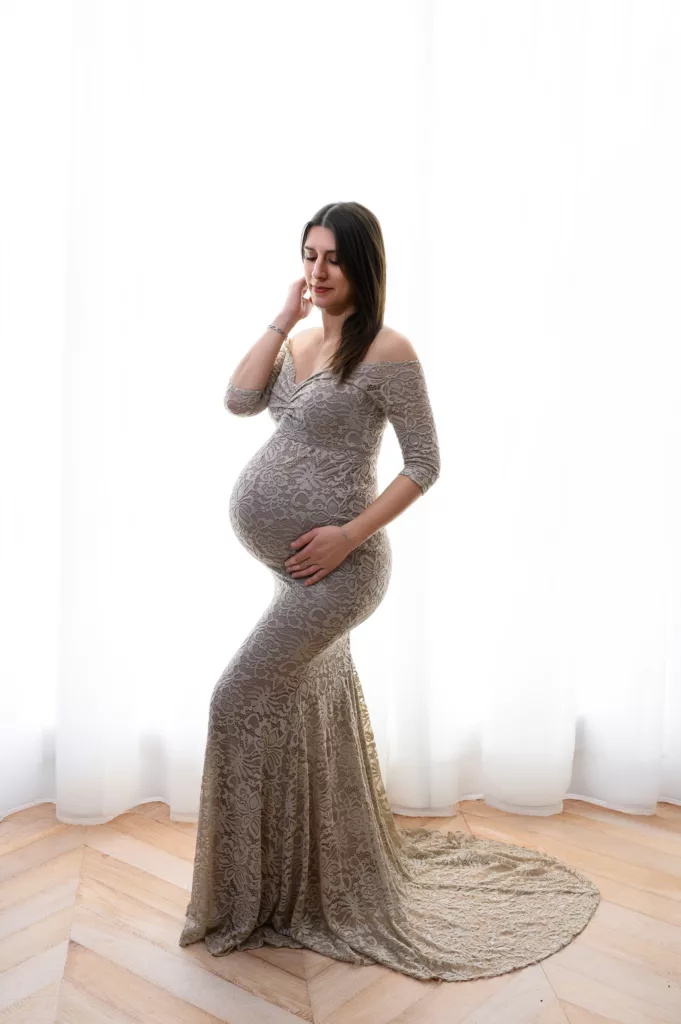 Pregnant woman in a long lace gown posing by sheer white curtains, hand on her belly.