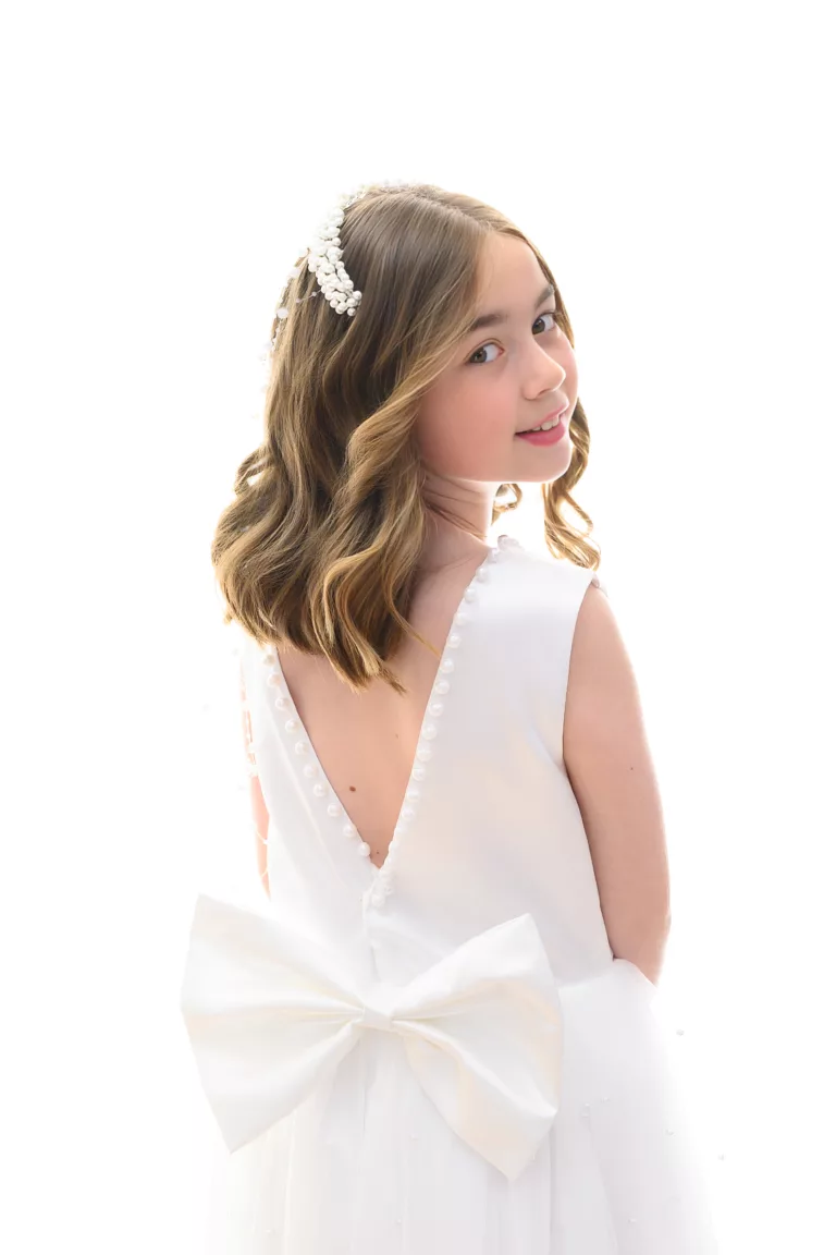 Young girl in a white dress with pearl trim, posing and looking over her shoulder.